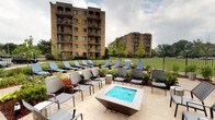 This outdoor amenity space is thoughtfully designed to serve as a communal retreat within a multi-building residential community. The centerpiece of the patio is a modern square fire-and-water feature finished in a dark metal casing, filled with vivid blue glass stones that create a striking visual focal point. Surrounding it, an arrangement of contemporary grey sling chairs and side tables provides comfortable seating for socializing or relaxing.

Extending outward from the central seating area, a row of blue cushioned sun loungers is positioned along a manicured landscaped border, offering dedicated spaces for sunbathing or unwinding in the open air. Ornamental shrubs, seasonal plantings in warm tones, and young trees add color and texture to the environment, creating a polished, resort-like atmosphere.

The patio surface is finished in neutral-toned pavers that complement the overall aesthetic. A wrought iron perimeter fence defines the space while maintaining open sightlines to a generous green lawn beyond. The backdrop features two multi-story buildings clad in warm brick, each with private balconies. The combination of structured landscaping, modern furnishings, and carefully chosen decorative elements gives the space an inviting and well-maintained character suited for outdoor leisure.