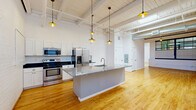 Located on University Avenue in Rochester, NY, this two-bedroom, one-bathroom loft-style unit blends industrial character with modern finishes across a generously sized single-floor layout. The space makes an immediate impression with soaring ceilings, exposed brick walls, and warm hardwood flooring that flows throughout the main living areas.

The open-concept kitchen and living area serves as the heart of the unit. The kitchen is well-appointed with stainless steel appliances — including a range, microwave, and French door refrigerator — white shaker cabinetry, dark countertops along the perimeter, and a large central island with a granite surface and built-in sink, illuminated by a row of pendant lights suspended from the exposed beam ceiling.

The primary bedroom offers a comfortable footprint with painted brick walls and exposed structural elements that maintain the loft aesthetic. The second bedroom mirrors this character, featuring hardwood floors and white-painted brick. A dedicated closet provides additional storage.

The bathroom is finished with large-format tile flooring, white cabinetry with granite countertops, and clean, neutral tones. A hallway connects the private rooms to the main living space. Steel-framed windows and visible ductwork complete the industrial-modern character that defines this distinctive Rochester residence.