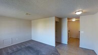 This single-floor space features a straightforward, practical layout designed for comfortable everyday living. The main living area is finished with wall-to-wall carpet in a neutral gray tone, providing a soft, understated foundation that pairs well with the plain white walls throughout. A textured popcorn ceiling runs across the entirety of the space, adding dimension overhead while housing a standard ceiling vent for climate control.

A partial dividing wall separates the carpeted living area from the adjacent hallway and utility zones, creating a natural transition between spaces. The hallway, visible in the background, leads to what appears to be a closet with a sliding door, offering storage access. Flooring shifts to tile in the kitchen and connecting areas, reflecting a practical material choice for higher-traffic and moisture-prone zones.

The kitchen area, partially visible to the right, features light-toned tile flooring and includes a stove, with warm overhead lighting that gives the space a cozy glow. The overall color palette is neutral and cohesive, utilizing beige, gray, and white tones throughout. The space maintains a clean, no-frills character suited to functional residential living.