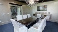 This single-story space showcases a refined interior centered around a dramatic meeting or dining room that blends contemporary design with upscale finishes. The room's focal point is a large, dark-lacquered rectangular table surrounded by plush, white upholstered chairs with skirted bases, offering both comfort and elegance. Overhead, a bold sputnik-style chandelier with crystalline metallic spires commands attention, complemented by a coffered ceiling with dark wood beam detailing that adds architectural depth and warmth. A wall-mounted flat-screen television is positioned above a built-in display ledge, which features decorative accents including a gold bowl and tiered ornamental pieces, reinforcing the room's polished aesthetic. The neutral taupe walls provide a calm, sophisticated backdrop, while large glass sliding doors along one side invite natural light and offer a visual connection to an adjacent space. Dark flooring grounds the room and contrasts beautifully with the lighter furnishings. Overall, the space exudes a contemporary yet warm ambiance, suited for formal dining, private meetings, or stylish entertaining.