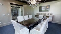 This single-story space showcases a refined interior centered around a dramatic meeting or dining room that blends contemporary design with upscale finishes. The room's focal point is a large, dark-lacquered rectangular table surrounded by plush, white upholstered chairs with skirted bases, offering both comfort and elegance. Overhead, a bold sputnik-style chandelier with crystalline metallic spires commands attention, complemented by a coffered ceiling with dark wood beam detailing that adds architectural depth and warmth. A wall-mounted flat-screen television is positioned above a built-in display ledge, which features decorative accents including a gold bowl and tiered ornamental pieces, reinforcing the room's polished aesthetic. The neutral taupe walls provide a calm, sophisticated backdrop, while large glass sliding doors along one side invite natural light and offer a visual connection to an adjacent space. Dark flooring grounds the room and contrasts beautifully with the lighter furnishings. Overall, the space exudes a contemporary yet warm ambiance, suited for formal dining, private meetings, or stylish entertaining.