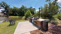 Located at 1500 Parklawn Dr in Charleston, South Carolina, this single-level outdoor amenity space is thoughtfully designed for community gathering and recreation. The centerpiece is a spacious covered gazebo-style pavilion with a classic white-painted wood railing and columns, a ceiling fan for warm Charleston days, and an open, airy design that invites abundant natural light and lush greenery views from every angle. The pavilion comfortably accommodates outdoor dining, furnished with a round patio table and vibrant orange chairs.

Adjacent to the pavilion, a dedicated outdoor grilling area features a built-in brick station equipped with multiple gas grills, ideal for community cookouts. This area overlooks a tranquil pond surrounded by mature trees, providing a serene natural backdrop. Seating alongside the grilling station allows residents to relax and enjoy the peaceful waterfront setting.

The exterior of the building is clad in warm yellow lap siding with crisp white trim and large double-hung windows, lending a welcoming, cottage-inspired aesthetic. Well-maintained landscaping with manicured shrubs lines the walkways. A nearby pool area enclosed by black iron fencing and accented by a Palmetto palm tree further highlights the Lowcountry character of this community amenity space.