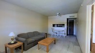 This single-floor space presents a straightforward and functional layout centered around an open living and dining area. The living room is furnished with a plush gray sofa accompanied by a wooden coffee table and a side table, creating a relaxed and inviting seating arrangement. A ceiling fan overhead provides both airflow and a subtle focal point to the room's neutral, light-colored walls and ceiling. Toward the back of the space, a compact dining area features a round white table with matching chairs, positioned adjacent to a built-in recessed nook that adds a unique architectural detail and provides additional utility. The flooring transitions from carpet in the living area to a harder surface near the entryway, adding visual definition between zones. A dark-toned entry door anchors the far end of the space, framed by the clean lines of the surrounding walls. The overall ambiance is calm and understated, with a neutral color palette throughout that lends itself well to a variety of uses. The space is practical in its design, offering an efficient and comfortable environment suited for everyday living.
