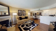 This well-appointed single-floor residence offers a thoughtfully designed open-concept layout that seamlessly connects the living, dining, and kitchen areas. The living room serves as the heart of the home, showcasing a tiled fireplace framed by a classic white mantle and a large decorative mirror above. A sliding glass door floods the space with natural light and provides direct access to an outdoor patio with views of the surrounding greenery. Warm hardwood-style flooring runs throughout, complementing the neutral, earthy tones of the walls and textured ceilings.

Adjacent to the living room, the dining area features a stylish tray ceiling detail and an elegant chandelier, creating an intimate setting for meals. Bold framed artwork adorns the walls, adding visual interest to the space.

The galley-style kitchen is efficiently laid out with crisp white cabinetry, ample counter space, recessed lighting, and a double stainless steel sink. It opens conveniently toward a utility passage at the rear.

The bedroom is a comfortable retreat, complete with natural window light, a spacious dresser area, and direct access to a well-lit closet. Two full bathrooms serve the residence, each featuring a tub-shower combination and clean white finishes, offering both functionality and simplicity.