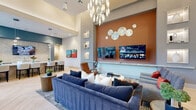 This well-appointed single-floor community lounge offers a generously sized, open-concept layout designed for both relaxation and social engagement. The space is anchored by a dramatic living area featuring soaring ceilings, a striking sculptural chandelier, and a bold terracotta accent wall adorned with decorative metallic disc art and a wall-mounted flat-screen television. Plush navy sofas and curated accent chairs create an inviting gathering space, complemented by built-in display niches showcasing decorative objects and vibrant artwork.

The herringbone-patterned light wood flooring flows seamlessly throughout, unifying the various zones of the lounge. A dedicated dining area offers round white tables paired with rich red chairs, set against teal accent walls and framed black-and-white artwork, with generous natural light streaming through floor-to-ceiling windows and sheer drapery.

A standout wet bar and café-style counter features an expansive marble surface with waterfall edges, upholstered bar seating, a tile backsplash, and pendant lighting overhead — ideal for hosting or casual gatherings. The entry area is framed by full-height glass doors and panels, providing transparency and a welcoming sense of arrival. Throughout the space, carefully selected furnishings, layered lighting, and curated art create a cohesive, sophisticated ambiance.