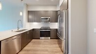 This 640-square-foot, one-bedroom, one-bathroom apartment at 1775 Metro Avenue in Chula Vista, California offers a thoughtfully designed single-floor layout. The open-concept kitchen and living area serves as the heart of the unit, featuring durable light-toned hardwood-style flooring throughout. The modern kitchen is equipped with dark wood cabinetry, stainless steel appliances including a gas range, microwave, and refrigerator, a white quartz peninsula countertop with pendant lighting overhead, and recessed ceiling lights. The living area is bright and airy, highlighted by large windows and a glass door that provide outdoor views and abundant natural light. The 130-square-foot bedroom features plush carpeting, a window with outdoor views, and direct access to two closets for ample storage. The full bathroom includes a shower/tub combination, a dark-toned vanity, built-in wall shelving, and hardwood-style flooring. Neutral gray and beige tones are used consistently throughout the unit, creating a clean, contemporary aesthetic. The well-organized floor plan maximizes the efficient use of space across all rooms.