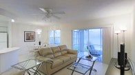This single-floor space showcases a seamless open-concept design that connects the living and dining areas in a light-filled, airy environment. The living area is anchored by a tan leather sofa and complemented by a glass-top coffee table, creating a relaxed yet polished atmosphere. Light-toned tile flooring runs throughout, enhancing the bright, clean aesthetic of the space.

Large windows and a sliding glass door flood the interior with abundant natural light while offering direct views of a screened outdoor patio, where additional seating is visible among lush tropical greenery. This indoor-outdoor connection gives the space a refreshing, open feel.

Overhead, a ceiling fan with an integrated light fixture provides both function and comfort. Adjacent to the living area, the dining space is distinguished by an elegant crystal chandelier that adds a touch of sophistication, set alongside a white dining table and chairs. Wall art provides a subtle decorative accent. Together, these elements create a cohesive, comfortable living environment with a bright, tropical character.