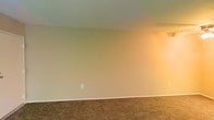 This single-story space presents a straightforward and functional interior designed to serve as a blank canvas for its occupants. The room visible features wall-to-wall carpeting in a warm tan hue, providing a soft underfoot feel that adds a degree of comfort to the space. The walls are finished in a neutral beige tone, creating a subdued and adaptable backdrop suitable for a variety of décor styles. White baseboards offer a clean contrast along the floor line, contributing to a tidy, finished appearance. The ceiling is smooth and painted in a crisp off-white, keeping the room feeling open and uncluttered. A single electrical outlet is centrally positioned along the main wall, indicating practical utility placement throughout the space. Warm ambient lighting, visible along one side of the room, lends a cozy atmosphere. The overall aesthetic is simple and unpretentious, emphasizing functionality and ease of customization. The space appears well-maintained and move-in ready, making it a practical option for those seeking a clean, neutral environment to personalize.