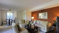 This single-floor living space offers a well-designed layout centered around a comfortable and stylishly furnished living room. The room is anchored by a neutral-toned sofa and coordinating armchairs, arranged around a dark wood coffee table adorned with decorative candles and greenery. A striking terracotta-orange accent wall serves as a bold focal point, complemented by a framed mirror that reflects light and adds visual depth to the space. A warm table lamp provides ambient lighting, enhancing the cozy atmosphere. The textured ceiling adds a classic architectural detail, while a visible ceiling vent indicates a functional climate control system. The entry door, fitted with a security viewer, is conveniently located within the living area, suggesting an open and accessible floor plan. Soft carpeting underfoot contributes to the comfortable feel of the room. The overall design blends warm tones with neutral furnishings, creating a balanced and inviting interior that feels both practical and aesthetically considered. Decorative accents, including a ceramic vase and candlestick holders, add personality and charm to the space.