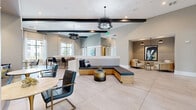 Located at 3030 Ascend Oak St in Apopka, Florida, this thoughtfully designed single-floor clubhouse offers a generous and versatile gathering space for residents. The entrance greets visitors with soaring ceilings, floor-to-ceiling black-framed glass doors, and expansive large-format tile flooring that flows seamlessly throughout the entire space, establishing an immediate sense of openness and sophistication.

The main lounge area is anchored by a built-in L-shaped bench with warm wood paneling, complemented by an assortment of sleek seating and striking statement chandeliers featuring dramatic feathered accents suspended from exposed dark ceiling beams. Exposed structural beams contrast beautifully against the neutral palette of warm beige and soft grey tones on the walls.

A café-style area provides bar-height counter seating along a bright white surface, flanked by tall windows that flood the space with natural light and offer views of the surrounding community. A cozy banquette nook nearby, framed in textured wall paneling, invites more intimate conversation or casual dining.

A dedicated alcove lounge offers a quieter retreat, accented by bold contemporary artwork and pendant lighting. Sliding barn doors open to reveal private individual workstations, each carpeted and decorated with vibrant botanical artwork, ideal for focused work or study. The overall ambiance is polished, welcoming, and distinctly modern.