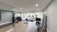 This fitness center is a thoughtfully designed, single-floor workout facility that offers a comprehensive range of exercise equipment within a clean, modern space. The interior features a neutral color palette of white and soft sage green, complemented by recessed lighting and large windows along one wall that flood the room with natural light and provide views of surrounding greenery.

The cardio zone is anchored by multiple Matrix-brand treadmills and an elliptical trainer, all positioned to face the outdoor scenery, enhancing the workout experience. A rowing machine rounds out the cardio offerings. For strength training, the space is equipped with a multi-functional cable machine, a leg press machine, a flat adjustable bench, and a full rack of dumbbells ranging across various weights. Floor-to-ceiling mirrors line one wall, visually expanding the space and allowing users to monitor their form during exercises.

The flooring transitions between rubberized fitness mats in the equipment zones and a speckled, durable composite surface, providing both safety and practicality. Framed athletic-themed artwork adds a motivational aesthetic touch to the walls. The layout is efficiently organized to maximize usability while maintaining clear circulation paths throughout. Overall, this fitness center delivers a professional, well-lit, and fully functional environment suitable for a wide range of workout routines.