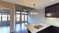 This single-floor residence showcases a thoughtfully designed open-concept layout that seamlessly connects the kitchen and living areas. The kitchen features dark cabinetry paired with speckled granite countertops and a stainless steel double-basin sink, with a stylish multi-bulb pendant light fixture hanging overhead to illuminate the workspace. Rich, dark hardwood flooring extends throughout the space, adding warmth and a polished aesthetic.

The living area benefits from an elevated exposed concrete ceiling detail, lending an industrial-modern character to the space. A ceiling fan is centrally mounted, providing both functionality and a finishing touch to the architectural ceiling. Natural light pours in generously through a set of French doors flanked by tall transom windows, which open onto what appears to be a private balcony with a railing, offering an outdoor extension of the living space and views of the surrounding landscape.

The neutral gray wall tones complement both the dark cabinetry and the concrete ceiling, creating a cohesive and contemporary color palette throughout. The overall ambiance is clean, bright, and modern, combining industrial design elements with refined finishes for a functional and visually appealing living environment.