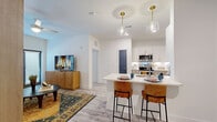This single-floor unit is designed with a contemporary, open-concept layout that seamlessly connects the living and kitchen areas. The kitchen is a standout feature, equipped with stainless steel appliances, two-tone cabinetry pairing white uppers with light wood-grain lowers, a white quartz waterfall island with an undermount sink, and seating for multiple bar stools. Brass pendant lights with glass shades add a refined touch overhead. Light wood-look flooring runs throughout, complementing the bright white walls and creating a cohesive aesthetic.

The living area is naturally lit by large windows with horizontal blinds and floor-length drapes, and includes a private entry door. A secondary sitting nook provides additional lounge space with a gallery wall arrangement.

The primary bedroom features large windows with a ceiling fan, ample natural light, and generous closet storage with wire shelving systems. A second bedroom makes a bold statement with a vibrant tropical tiger-print accent wall, navy upholstered headboard, and warm amber textiles. The bathroom includes a full soaking tub and shower combination with large-format tile surround. Throughout, the design balances modern finishes with warm, eclectic décor details.