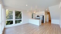 This contemporary two-floor space is designed with a clean, modern aesthetic that emphasizes openness and natural light. The main living area flows seamlessly into the kitchen, creating an open-concept layout ideal for everyday living and entertaining. Light-toned hardwood flooring runs throughout, lending warmth and continuity to the space.

The kitchen is well-appointed with white shaker-style cabinetry, stainless steel appliances including a refrigerator, microwave, and range, and a central island with a sink that doubles as a breakfast bar. Elegant pendant lighting hangs above the island, adding a refined touch to the functional workspace.

Large windows frame lush, tree-lined views, filling the living area with generous natural light and creating a bright, airy atmosphere. Recessed lighting throughout the ceiling supplements the natural light, ensuring the space is well-illuminated at any time of day.

The overall design palette is neutral and fresh, with white walls, crisp trim, and light finishes that give the space a clean, move-in-ready feel. The second floor offers additional living space, extending the functionality of this thoughtfully designed property. The combination of modern finishes, open layout, and verdant outdoor views makes this a bright and inviting space.