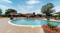 Located at 1870 Enterprise Drive in Virginia Beach, Virginia, this single-floor community facility offers a well-appointed layout designed for resident amenity and leisure. The entry corridor sets a welcoming tone with warm cream-colored walls, rich multi-toned slate tile flooring, and coastal-themed artwork — including framed photography of sailboats and waterfront scenes — that nod to the region's maritime character. The hallway provides access to clearly marked gender-separated restrooms and connects directly to the main gathering space through a glass-paneled door, allowing natural light to flow through the interior. The main room, visible through the glass partition, features tile flooring, ceiling fans, and a bright, open ambiance with tasteful furnishings. A fire alarm system is in place, underscoring the building's attention to safety. The exterior showcases the facility's standout amenity: a generously sized freeform outdoor pool framed by a paved surround, lounge chairs, and umbrella-shaded seating areas. Mature trees provide shade along the perimeter, while colorful plantings add seasonal vibrancy. The pool area is complemented by an adjacent poolhouse structure, creating an inviting outdoor retreat suited for warm Virginia Beach summers.
