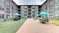 This expansive open-air courtyard is nestled within the interior of a multi-story residential building, providing a sheltered yet open-sky environment for outdoor recreation and relaxation. The courtyard is designed with distinct functional zones that cater to a variety of activities.

A lush turf lawn serves as the central feature, anchored by multiple freestanding hammocks on arched wooden frames, ideal for lounging. Surrounding the lawn, a brick-paver walkway connects the various amenity areas, creating a cohesive flow throughout the space. Wicker lounge chairs with deep navy cushions are grouped beneath large teal market umbrellas, offering shaded seating areas.

A dedicated fire pit zone features a square gas fire table surrounded by vibrant teal Adirondack chairs, providing an inviting gathering spot. Adjacent to this area, a built-in outdoor grilling station with a stainless steel gas grill and stone countertops offers a practical space for outdoor cooking, complemented by nearby dining tables and chairs.

Decorative landscaping, including ornamental grasses, shrubs, and a striking oversized ceramic planter, adds visual interest throughout. The surrounding multi-story building facade, with its balconies and large windows, frames the courtyard and contributes to its enclosed, community-oriented ambiance.