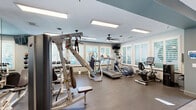 Located at 3301 Arena Blvd in Sacramento, California, this fitness center offers a thoughtfully designed exercise space that caters to a wide range of workout needs. The facility is divided into two distinct zones: a rubber-floored workout area and a light wood-floored open space, ideal for stretching, yoga, or functional movement exercises.

The main workout area is lined with plantation-shuttered windows that allow ample natural light to filter in while maintaining privacy. Full-length mirrors along one wall visually expand the space and assist with form during exercise. The room is equipped with a comprehensive selection of cardio machines, including a treadmill, elliptical, and stationary bike, as well as a multi-function cable machine, adjustable weight bench, a full dumbbell rack, and medicine balls. Ceiling fans provide additional ventilation, and a wall-mounted TV keeps users entertained during workouts.

The adjacent open area features light hardwood-style flooring, recessed lighting, and teal acoustic panels that add a modern aesthetic while enhancing sound quality. Framed artwork and clean, neutral walls contribute to a calm and welcoming atmosphere. Together, both zones create a versatile, well-lit fitness environment designed to support a variety of exercise routines.