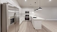 This two-story residence is thoughtfully laid out across two levels, with the main living areas occupying the upper floor and a compact entry level below connected by a carpeted staircase with a sleek metal handrail. The open-concept kitchen and living room form the heart of the home, showcasing white shaker cabinetry, stainless steel appliances including a refrigerator, range, and microwave, and a peninsula countertop finished in light granite with an undermount sink. Stylish black pendant lights hang above the peninsula, adding a modern touch, while light wood-look flooring runs throughout this level, creating a clean and cohesive aesthetic. The neutral-toned living area flows naturally from the kitchen, benefiting from natural light through windows dressed with blinds. The spacious primary bedroom features plush carpeting, a ceiling fan, a natural light-admitting window, and direct access to a dedicated closet. The full bathroom includes a tub-shower combo, granite-top vanity, and wood-look flooring. A dedicated laundry room with a full-size washer and dryer and overhead wire shelving adds everyday convenience. The space also includes a separate closet for additional storage.
