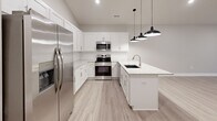 This two-story residence is thoughtfully laid out across two levels, with the main living areas occupying the upper floor and a compact entry level below connected by a carpeted staircase with a sleek metal handrail. The open-concept kitchen and living room form the heart of the home, showcasing white shaker cabinetry, stainless steel appliances including a refrigerator, range, and microwave, and a peninsula countertop finished in light granite with an undermount sink. Stylish black pendant lights hang above the peninsula, adding a modern touch, while light wood-look flooring runs throughout this level, creating a clean and cohesive aesthetic. The neutral-toned living area flows naturally from the kitchen, benefiting from natural light through windows dressed with blinds. The spacious primary bedroom features plush carpeting, a ceiling fan, a natural light-admitting window, and direct access to a dedicated closet. The full bathroom includes a tub-shower combo, granite-top vanity, and wood-look flooring. A dedicated laundry room with a full-size washer and dryer and overhead wire shelving adds everyday convenience. The space also includes a separate closet for additional storage.