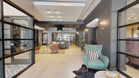 Located at 346–358 Trumbull Street in Hartford, CT, this single-floor hallway space serves as a thoughtfully designed lobby entrance. The layout flows from a dramatic glass-and-steel entry vestibule, where floor-to-ceiling black-framed glazing floods the space with natural light and offers clear views to the streetscape beyond. Flanking the entry are manicured topiary plants, reinforcing a polished, welcoming aesthetic.

The main lounge area is generously proportioned, anchored by a bold teal-tiled fireplace wall fitted with a wall-mounted television and flanked by built-in open shelving displaying decorative objects. Comfortable seating arrangements—featuring a large sectional sofa, mid-century-style accent chairs in warm terracotta tones, and a teal wingback chair—are organized atop a striking patterned area rug, creating distinct conversation zones. Brass wall sconces and a dramatic oversized pendant light contribute to the layered, contemporary lighting scheme.

Adjacent to the lounge, a dedicated package and mail room houses a wall of individual mailboxes alongside a bank of secure parcel lockers, accented by colorful abstract artwork. The continuous large-format tile flooring and a tiered ceiling with recessed lighting unify the entire hallway, lending the space a cohesive, upscale ambiance throughout.