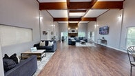 Located at 750 Gaines School Rd in Athens, Georgia, this single-floor space offers a thoughtfully designed layout that balances openness with functionality. The standout feature of the living room is its dramatic vaulted ceiling with exposed wooden beams, creating an airy, expansive atmosphere. Rich hardwood-style flooring runs throughout, complementing the neutral gray walls. A full-height brick fireplace with a solid wood mantel serves as the living room's focal point, adding warmth and character. Natural light filters in through multiple windows fitted with blinds, offering views of the surrounding greenery.

The kitchen is efficiently designed with white shaker-style cabinetry extending to the ceiling, providing ample storage. A granite-style countertop lines the workspace, which includes a stainless steel sink, black appliances, and under-cabinet lighting. A pass-through bar counter opens the kitchen to the adjoining area, where bar stools create a casual dining or gathering space that enhances the open-concept feel.

A connecting hallway provides a transitional space between the main rooms, contributing to the smooth flow of the overall floor plan. Together, the rooms create a cohesive, welcoming environment suited for both everyday living and entertaining.