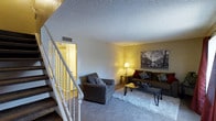 This two-story residential space offers a functional layout spread across two levels, connected by a staircase with a classic white railing visible from the main living area. The ground floor features a welcoming living room with plush carpeting, a textured ceiling, and warm ambient lighting that creates a cozy atmosphere. The staircase anchors one side of the room, contributing to the sense of vertical space and openness within the home.

The living area is generously sized, providing ample room for seating arrangements and everyday living. Natural and artificial lighting work together to illuminate the interiors, with warm-toned walls adding to the inviting character of the space.

Additional areas of the home reveal wood-style flooring and evidence of active renovation work, including exposed ceiling structures, removed drywall sections, and visible HVAC components. These areas indicate that portions of the space are currently mid-renovation, offering future occupants or investors the opportunity to see the home's underlying infrastructure and potential.

A bathroom is partially visible during the renovation phase, showing a toilet and sink fixtures in place. Overall, this space presents a combination of move-in-ready living areas alongside sections undergoing significant improvement, making it a property with clear renovation and customization potential.