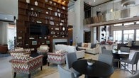 This stylish communal lounge space makes an immediate impression with its soaring double-height ceilings and an open, airy atmosphere. The focal point of the room is a floor-to-ceiling dark wood built-in shelving unit, artfully styled with decorative objects, books, and greenery, flanking a contemporary fireplace with a sleek surround. A large flat-screen television is integrated seamlessly into the shelving arrangement, adding a functional yet refined touch.

The seating area is thoughtfully arranged to encourage both conversation and relaxation, featuring a mix of bold patterned accent chairs in warm earth tones alongside neutral upholstered sofas and armchairs. This blend of textures and colors creates an inviting, curated ambiance. A round black occasional table in the foreground adds contrast and anchors the space visually.

An upper-level mezzanine with glass and metal railing is visible above, contributing to the dramatic vertical scale of the interior. Recessed lighting and track lighting illuminate the space evenly, complementing the modern, sophisticated design aesthetic. The light-toned flooring and neutral walls provide a clean backdrop that allows the rich furnishings and architectural details to stand out, resulting in a space that is both welcoming and visually dynamic.