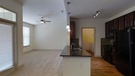 This single-floor space showcases a thoughtfully designed open-concept layout that connects the kitchen and living areas in a cohesive, functional arrangement. The kitchen is a standout feature, boasting a large peninsula with striking dark granite countertops and an undermount sink, making it ideal for both meal preparation and casual dining. Pendant lights hang above the peninsula, adding a warm, focused illumination, while a track lighting system overhead provides additional brightness. Dark-stained cabinetry lines the kitchen walls, offering generous storage and lending a rich contrast to the neutral-toned surroundings. A tile backsplash adds texture and visual interest behind the cooking area. The kitchen flows naturally into the living area, which is finished with plush wall-to-wall carpeting, creating a comfortable and inviting atmosphere. A ceiling fan with an integrated light fixture anchors the living space, ensuring both comfort and adequate lighting throughout. The clean, light-colored walls throughout the space contribute to an airy, open feel. A glimpse through a doorway reveals an additional room beyond the kitchen, hinting at further utility within the home. The overall ambiance is modern, clean, and well-appointed.