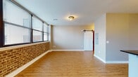 This thoughtfully designed single-floor unit seamlessly combines industrial character with modern finishes. The open-concept living area is anchored by warm hardwood-style flooring and a striking exposed brick accent wall, with large windows fitted with blinds that allow natural light to fill the space while offering city views. A sliding barn door adds a distinctive architectural touch, connecting the living area to adjoining spaces.

The kitchen is a standout feature, offering white shaker cabinetry with matte black hardware, dark granite countertops, a white subway tile backsplash, and stainless steel appliances including an electric range and over-the-range microwave. Open reclaimed wood shelving suspended from black iron rods provides both practical storage and an industrial aesthetic. A peninsula with additional counter space and a matte black faucet enhances functionality.

The bedroom is generously proportioned with neutral grey walls and benefits from ample storage courtesy of two side-by-side reach-in closets, each outfitted with wire shelving and hanging rods. The bathroom features a tiled walk-in shower enclosed by a sleek black-framed frosted glass door, complemented by coordinating black hardware. Throughout the unit, consistent flooring, clean trim, and a cohesive neutral color palette create a polished and inviting atmosphere.