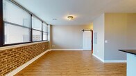 This thoughtfully designed single-floor unit seamlessly combines industrial character with modern finishes. The open-concept living area is anchored by warm hardwood-style flooring and a striking exposed brick accent wall, with large windows fitted with blinds that allow natural light to fill the space while offering city views. A sliding barn door adds a distinctive architectural touch, connecting the living area to adjoining spaces.

The kitchen is a standout feature, offering white shaker cabinetry with matte black hardware, dark granite countertops, a white subway tile backsplash, and stainless steel appliances including an electric range and over-the-range microwave. Open reclaimed wood shelving suspended from black iron rods provides both practical storage and an industrial aesthetic. A peninsula with additional counter space and a matte black faucet enhances functionality.

The bedroom is generously proportioned with neutral grey walls and benefits from ample storage courtesy of two side-by-side reach-in closets, each outfitted with wire shelving and hanging rods. The bathroom features a tiled walk-in shower enclosed by a sleek black-framed frosted glass door, complemented by coordinating black hardware. Throughout the unit, consistent flooring, clean trim, and a cohesive neutral color palette create a polished and inviting atmosphere.