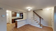 This two-story space offers a practical and well-appointed layout, beginning with an open-concept main level that seamlessly integrates the kitchen and living areas. The kitchen is outfitted with rich, dark wood cabinetry that provides ample storage, complemented by light countertops that create a pleasing contrast. A peninsula-style breakfast bar extends from the kitchen, offering additional workspace and a casual dining option. Black appliances, including a refrigerator and range, are neatly integrated into the cabinetry. A stainless steel sink is positioned beneath a window, allowing for natural light in the kitchen area. The main level features warm hardwood-style flooring that runs throughout, adding a cohesive and inviting feel to the space. A flush-mount ceiling light provides ambient illumination. A white-paneled door on the main floor leads to an additional room or storage area. The staircase, featuring a classic wooden banister with white spindles and natural wood treads, leads to the second floor, adding an architectural element that draws the eye upward. The neutral wall tones throughout create a clean, versatile backdrop suitable for a variety of interior styles.