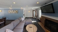 This single-floor community room is thoughtfully designed to serve as a versatile shared gathering space. The lounge area anchors the room with a comfortable sectional sofa, a round wood coffee table, and a wall-mounted flat-screen television positioned above a tile-surround fireplace. Recessed lighting and large windows allow natural light to fill the space, complementing the cool blue-gray wall tones and dark wood-plank flooring that run throughout.

Adjacent to the lounge, a dedicated dining area accommodates a dark wood table with cushioned chairs, accented by a wall sconce and framed artwork. The layout flows openly between the seating and dining zones, making the space well-suited for both casual socializing and organized events.

The fully equipped kitchen features warm wood cabinetry paired with gray countertops, a double-basin sink, and stainless steel appliances including a refrigerator, electric range, over-the-range microwave, and dishwasher. Pendant lighting and a vibrant piece of wall art add a modern touch. A side window brings in additional natural light, enhancing the functionality and atmosphere of the kitchen. Together, these areas create an inviting, practical community space ideal for resident use and group gatherings.