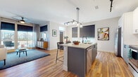 This well-appointed single-floor residence showcases a thoughtfully designed open-concept layout with warm hardwood flooring flowing throughout the main living areas. The kitchen serves as a central hub, featuring a large island with a light quartz countertop, integrated sink, stainless steel appliances including a French door refrigerator and range, white cabinetry with a tile backsplash, and stylish pendant lighting overhead. The island offers bar seating, making it ideal for casual dining or entertaining. The living room is bathed in natural light from floor-to-ceiling windows and sliding glass doors that open to a private balcony, offering open outdoor views. The space comfortably accommodates a full seating arrangement anchored by rich, colorful artwork on the walls. The primary bedroom features plush carpeting, large windows with roller shades and drape panels, and a distinctive arched wall detail. A second bedroom also features carpeted floors and direct access to a closet. The primary bathroom is finished with large-format tile flooring, a wide white vanity with black hardware, a glass-enclosed shower, and a soaking tub. A second full bathroom mirrors the same clean aesthetic, featuring a tub-shower combination and matching tile finishes throughout.