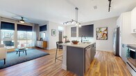 This well-appointed single-floor residence showcases a thoughtfully designed open-concept layout with warm hardwood flooring flowing throughout the main living areas. The kitchen serves as a central hub, featuring a large island with a light quartz countertop, integrated sink, stainless steel appliances including a French door refrigerator and range, white cabinetry with a tile backsplash, and stylish pendant lighting overhead. The island offers bar seating, making it ideal for casual dining or entertaining. The living room is bathed in natural light from floor-to-ceiling windows and sliding glass doors that open to a private balcony, offering open outdoor views. The space comfortably accommodates a full seating arrangement anchored by rich, colorful artwork on the walls. The primary bedroom features plush carpeting, large windows with roller shades and drape panels, and a distinctive arched wall detail. A second bedroom also features carpeted floors and direct access to a closet. The primary bathroom is finished with large-format tile flooring, a wide white vanity with black hardware, a glass-enclosed shower, and a soaking tub. A second full bathroom mirrors the same clean aesthetic, featuring a tub-shower combination and matching tile finishes throughout.