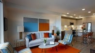 This single-floor space offers a thoughtfully designed open-plan layout that blends style and functionality. The living area is a standout, anchored by a light gray sofa adorned with bold orange and blue accent pillows, complemented by a striped multicolor lounge chair that adds a playful, retro-inspired touch. An oval white coffee table with sleek hairpin legs sits atop a vibrant orange area rug, tying the color palette together. Two large abstract artworks — featuring blue and orange color blocks — hang prominently on the wall, reinforcing the space's energetic aesthetic. Recessed lighting overhead ensures the area is well-lit, while the white walls keep the overall feel fresh and airy. The living space flows seamlessly into a contemporary kitchen, which features clean-lined cabinetry in a warm neutral tone, stainless steel appliances including a microwave and dishwasher, and a compact countertop workspace with under-cabinet lighting. The open connection between the kitchen and living area creates a sense of spaciousness and promotes easy interaction across the floor plan. The overall ambiance is modern and lively, with a carefully curated mix of color and texture throughout.