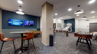 This thoughtfully designed community game room offers an engaging recreational space spread across a single floor. The interior features a contemporary aesthetic, with bold teal accent walls contrasting against neutral beige tones and crisp white ceilings. Patterned, multi-toned carpet in shades of grey and charcoal runs throughout, lending texture and visual interest underfoot.

The space is generously equipped with a variety of entertainment options. A full-length wooden shuffleboard table anchors one side of the room, while a foosball table provides additional competitive fun nearby. An air hockey table and a basketball shooting arcade game round out the gaming offerings, catering to a wide range of interests. Structural columns are integrated seamlessly into the layout without interrupting the open, airy feel of the room.

A dedicated lounge and dining area features sleek pedestal tables paired with cognac leather chairs, positioned beneath wall-mounted flat-screen TVs. Stylish pendant lights and recessed ceiling fixtures provide warm, layered illumination throughout. A wet bar with dark cabinetry, a countertop sink, and open shelving offers a convenient refreshment station.

Curated artwork, including framed prints and abstract canvases, is displayed throughout, adding a polished, gallery-like character to the space. The overall ambiance is modern, social, and inviting, making it an ideal setting for both casual gatherings and competitive play.