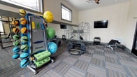 This single-floor fitness space is thoughtfully designed to support a wide variety of workout routines. The room is outfitted with an impressive selection of equipment, including a Smart Fitness equipment rack stocked with stability balls in multiple sizes, resistance bands, and foam rollers for mobility and recovery work. A functional trainer frame provides options for bodyweight and suspension training, while a set of kettlebells on a dedicated rack caters to strength and conditioning exercises. A balance board adds a dynamic training option to the mix. A wall-mounted flat-screen television keeps users entertained or connected to guided workout content during sessions. The room is finished with durable patterned carpet tiles that add texture and visual interest underfoot. Natural light filters in through multiple windows positioned near the ceiling, brightening the space and creating an open, energizing atmosphere. A ceiling fan ensures comfortable airflow during intense workouts. The neutral-toned walls and practical layout allow the equipment to remain the focal point, making this a functional and inviting environment for fitness enthusiasts of all levels.