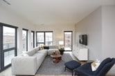 This single-floor residence showcases a thoughtfully designed open-concept layout with a seamless flow between the living, dining, and kitchen areas. The living room is a bright, welcoming space anchored by a generous sectional sofa and a trio of black-framed windows offering urban views. Light wood flooring runs throughout the main living areas, complementing the neutral, modern color palette.

The kitchen is well-equipped with two-tone cabinetry featuring white upper cabinets and warm wood-grain lower cabinets, crisp white quartz countertops, a subway tile backsplash, and a full suite of stainless steel appliances. A central island provides additional prep space and casual seating. Track lighting overhead adds a contemporary touch.

The residence includes multiple bedrooms, each carpeted and finished in soft neutral tones. One bedroom features a ceiling fan and framed botanical artwork, while another includes large windows with natural light and a stylish six-drawer dresser. Both rooms offer comfortable proportions and a calm ambiance.

The bathrooms are finished with large-format grey tile, with one bathroom offering a tub-shower combination and another featuring a dedicated walk-in shower with chrome fixtures. Overall, the space presents a clean, contemporary aesthetic with functional design throughout.