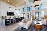Located at 4660 Derrick Rd in Union City, Georgia, this single-floor community clubhouse delivers a sophisticated and welcoming atmosphere through thoughtful architectural design and upscale finishes. The space is defined by dramatic double-height ceilings that flood the interior with natural light through expansive clerestory windows arranged along the upper walls, creating an airy, open feel throughout.

The main lounge area serves as the social centerpiece, featuring plush neutral seating, a large-format wall-mounted television, and designer pendant lighting that anchors the room with modern elegance. Large-format tile flooring transitions seamlessly between zones, while bold patterned area rugs delineate comfortable conversation and relaxation areas.

A standout bar and counter area is lined with stylish upholstered barstools set against a striking geometric patterned accent wall that extends to the full height of the ceiling. A white quartz countertop island, integrated sink, and warm wood cabinetry add a refined, contemporary feel to this gathering spot. Pendant lights with a wood-paneled ceiling inset provide a warm contrast to the crisp white walls.

The billiards area offers a full-size pool table beneath bold drum pendant lights, with a wall-mounted TV and direct sightlines to the outdoor pool area through glass doors. Slatted wood accent panels add warmth and texture throughout the space, tying together the modern and natural design elements cohesively.
