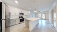 This well-designed 1-bedroom, 1-bathroom unit offers a clean, modern aesthetic across a single floor. Polished concrete floors run throughout the entire space, providing a sleek, low-maintenance foundation that reflects natural light effectively. The open-concept kitchen and living area serves as the heart of the unit, anchored by a large central island with a built-in sink and pendant lighting overhead. The kitchen is equipped with stainless steel appliances, including an electric range and over-the-range microwave, complemented by white shaker-style cabinetry with gold-toned hardware and bright white countertops. The living area extends toward a glass exterior door and windows, allowing ample natural light and outdoor views. A ceiling fan adds to the comfort of this space. The bedroom is a comfortable size, featuring a ceiling fan, natural light from a window with blinds, and direct access to the walk-in closet, which offers multi-tiered shelving and hanging rods for organized storage. The bathroom features a long vanity with white countertops, undermount sink, gold hardware accents, and a backlit mirror, maintaining the unit's consistent modern design language throughout.