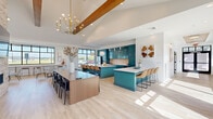 Located at 249 Allure Ln in Chesapeake, Virginia, this expansive single-floor amenity space offers a thoughtfully designed interior that blends modern aesthetics with functional, community-focused living. The open-concept kitchen and living area serves as the heart of the space, featuring striking teal cabinetry that extends floor to ceiling, a warm terracotta tile backsplash, and gleaming white quartz countertops across dual islands — one serving as a prep and seating bar, the other as a large communal dining table. Exposed wood beams arc across the vaulted ceiling, adding rustic warmth against bright white walls, while a statement brass chandelier anchors the dining area. Light hardwood flooring flows throughout, enhancing the airy, open feel. Large black-framed windows flood the interior with natural light and offer views of the surrounding greenery. The living area showcases a floor-to-ceiling stacked stone fireplace with a mounted television, complemented by an adjacent recreation lounge equipped with a billiards table and comfortable seating. A dedicated home theater room provides a complete cinematic experience, featuring tiered rows of leather reclining seats, a large projection screen, a ceiling-mounted projector, and dark, acoustically treated walls for an immersive atmosphere.