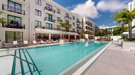 Located at 24 NE First Ave in Dania Beach, Florida, this outdoor amenity level is designed to deliver a resort-style experience within a modern multi-story residential community. The centerpiece of the space is a generously sized swimming pool with crystal-clear turquoise water, framed by wide white porcelain tile decking and lined with lounge chairs ideal for sunbathing. Tall, mature palm trees are strategically placed throughout the courtyard, providing natural shade and reinforcing the tropical South Florida aesthetic. Large raised planter beds with lush tropical foliage serve as elegant landscaping dividers throughout the pool deck. A dedicated sun lounge area features shaded cabana structures set over manicured artificial turf, offering a comfortable retreat from the sun. The surrounding multi-story building displays a clean, contemporary white façade with black-railed balconies and floor-to-ceiling glass doors at the ground level, creating a seamless visual connection between interior and exterior spaces. Wooden-treaded staircases with sleek dark metal railings provide access between levels, blending warmth with modern design. The overall ambiance is bright, open, and vacation-inspired, making this amenity level a defining feature of the Dania Beach property.