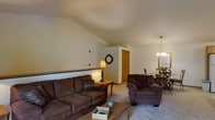 This single-floor residence offers a welcoming and functional layout highlighted by a standout vaulted ceiling in the main living area. The angled ceiling adds a sense of vertical openness to the space, creating an airy atmosphere despite the cozy, intimate feel of the room. The living area is carpeted and centered around plush, dark brown upholstered seating, including a sofa and armchair, arranged around a wooden coffee table. A small side table with a warm lamp adds ambient lighting to the corner of the room. The walls are finished in soft, neutral tones that complement the warm-toned furnishings throughout. A low half-wall or ledge divides the living area from the adjacent dining space, which is visible in the background and features a chandelier overhead, providing elegant accent lighting. The dining area accommodates a table and chairs, making it well-suited for everyday meals. A bi-fold closet door is visible near the dining space, offering convenient storage. The overall ambiance of the home is warm and understated, with a traditional aesthetic that prioritizes comfort and practicality in a straightforward single-level floor plan.