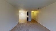 This single-floor residence offers a straightforward and functional layout designed for comfortable everyday living. The main living area is finished with neutral wall-to-wall carpeting, creating a warm and cohesive feel throughout the space. A textured ceiling adds a classic touch overhead, while low-profile baseboard heating lines the perimeter of the room.

The living space opens directly into a dining area, distinguished by a modest chandelier that provides a focal point and adds a touch of elegance. Adjacent to the dining area, the kitchen features dark cabinetry that provides a visual contrast against the lighter walls and flooring. The kitchen area transitions to a tile or vinyl floor, offering a practical surface suited for food preparation.

A hallway connects to additional areas of the home, with a door visible at the far end suggesting access to a bedroom or utility space. The layout flows naturally from room to room, making the most of the available footprint. Throughout the space, recessed and surface-mounted ceiling lights provide consistent illumination. The overall ambiance is clean, neutral, and move-in ready, offering a blank canvas well-suited for personalization.