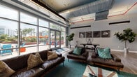 Located at 100 Commons Park N in Stamford, Connecticut, this single-floor amenity space blends industrial-chic design with comfortable, contemporary furnishings throughout. The main lounge area is a standout, featuring exposed ductwork and vivid red piping against a high ceiling, floor-to-ceiling windows, and large glass doors that flood the space with natural light and open directly to an outdoor terrace. Rich brown leather sofas, teal accent rugs including a bold Union Jack design, and lush fiddle-leaf fig plants create a welcoming and stylish atmosphere. A sleek, linear fireplace set into a textured white wall panel serves as a dramatic focal point in the adjacent seating area, complemented by a wall-mounted television and game table with industrial stools. The kitchen features a generous island with reclaimed wood barstools, white subway tile backsplash, stainless steel appliances, and decorative bicycle wall art. Conference rooms are finished in calming blue tones, adorned with framed butterfly artwork, and furnished with solid wood tables on casters and leather chairs — ideal for focused meetings. Additional seating areas with expansive windows offer serene tree-lined views, making this a versatile, thoughtfully designed community space.