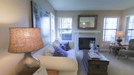 This single-floor residence offers a thoughtfully designed living space that balances comfort and style. The living room serves as the heart of the home, anchored by a classic brick fireplace with a clean white mantel, creating a warm and inviting focal point. An ornate, decoratively framed mirror above the mantel adds an elegant touch to the space. The room is furnished with a plush white sofa adorned with soft accent pillows in neutral and muted tones, complemented by a rich, dark wood coffee chest that provides both storage and character. A textured table lamp adds warmth to the ambiance. Large windows along one wall allow generous natural light to pour in, offering views of lush outdoor greenery and creating a bright, airy atmosphere throughout the day. The walls are finished in soft, warm neutral tones that contribute to a calm and cohesive aesthetic. A textured ceiling adds subtle architectural interest overhead. The overall ambiance of the space is relaxed yet refined, with carefully chosen décor elements — including framed wall art depicting a nature scene — that enhance the sense of a welcoming, well-curated home environment.