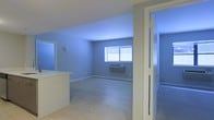 This single-floor space offers a clean, minimalist layout designed for efficient everyday living. The main living area is finished with light wood laminate flooring that runs throughout, contributing to a bright and airy feel. Soft blue-toned walls add a calm, neutral character to the interior, while white trim and baseboards provide a crisp contrast throughout.

A window along the far wall of the living area allows natural light to filter in, illuminating the open floor plan. A wall-mounted HVAC unit beneath the window provides climate control directly within the living space. The kitchen area features a raised counter or breakfast bar that visually separates it from the main living zone while maintaining an open connection between the two areas.

To one side, a bathroom is accessible through a white-framed door with frosted glass panel insets, offering privacy while maintaining a modern aesthetic. The bathroom, visible from the entryway, appears to include standard fixtures and continues the clean, functional design language found throughout the rest of the space.

Overall, the space presents a straightforward, well-maintained interior that prioritizes functionality and simplicity, making it a practical and comfortable single-floor living environment.