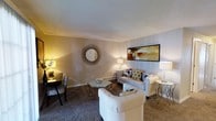 This single-floor residence features a 2-bedroom, 1-bathroom layout designed for comfortable everyday living. The living room serves as the heart of the home, showcasing a warm, neutral color palette with soft beige walls and plush wall-to-wall carpeting that adds a cozy feel underfoot. The space is thoughtfully arranged to accommodate both relaxing and casual dining, with room for a sofa, accent chairs, and a dining set tucked neatly to the side. A decorative sunburst mirror and framed wall art add personality and visual interest to the living area, while multiple floor and table lamps provide warm ambient lighting throughout. The textured ceiling adds a classic touch to the overall aesthetic. Baseboard heating runs along the walls, ensuring year-round comfort. The open flow of the living space allows for versatile furniture arrangements, making it easy to personalize. The neutral tones and soft finishes create a calm, inviting ambiance that carries throughout the home, making this a practical and pleasant place to live.