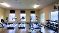 This fitness center is designed to provide a functional and comfortable workout environment. The space is outfitted with a variety of cardio equipment, including multiple treadmills, elliptical machines, and a rowing machine, offering users a well-rounded selection of exercise options. Several flat-screen TVs are mounted on the walls, allowing members to stay entertained during their workouts. The room benefits from generous natural light, courtesy of multiple large windows that line the walls and offer views of the surrounding outdoor area, creating an open and energizing atmosphere. The flooring is a durable hard surface suited for high-traffic fitness use, and the warm, neutral-toned walls contribute to the space's clean and welcoming feel. A ceiling fan provides additional air circulation, complementing the overhead fluorescent lighting that ensures the room is well-lit at all times. The single-story layout keeps everything conveniently accessible, and the overall design prioritizes practicality while maintaining a pleasant, motivating environment for users of all fitness levels.