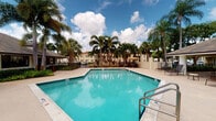 This single-floor amenity space in Miami, FL offers residents a comprehensive fitness and recreation experience in a well-maintained tropical setting. The fitness center is a generously sized, bright space designed with functionality in mind. Its white walls, recessed lighting, and rubber flooring create a clean and practical workout environment. Large windows and glass double doors on multiple sides flood the room with natural light and provide views of the surrounding greenery and parking areas. The gym is outfitted with a wide variety of equipment, including treadmills, stair climbers, stationary bikes, and an extensive selection of strength-training machines to support both cardio and resistance training routines. The layout is thoughtfully organized to allow comfortable movement throughout the space. Outside, the resort-style swimming pool is a standout feature, offering a spacious, sparkling turquoise pool framed by tall swaying palm trees and lush tropical foliage. The pool deck provides ample room for lounging, with seating areas and shaded spots nearby. The backdrop of the multi-story building behind the pool, set against the characteristically bright blue Miami sky, reinforces the tropical ambiance. Together, these amenities deliver a well-rounded recreational experience suited to an active lifestyle in South Florida.